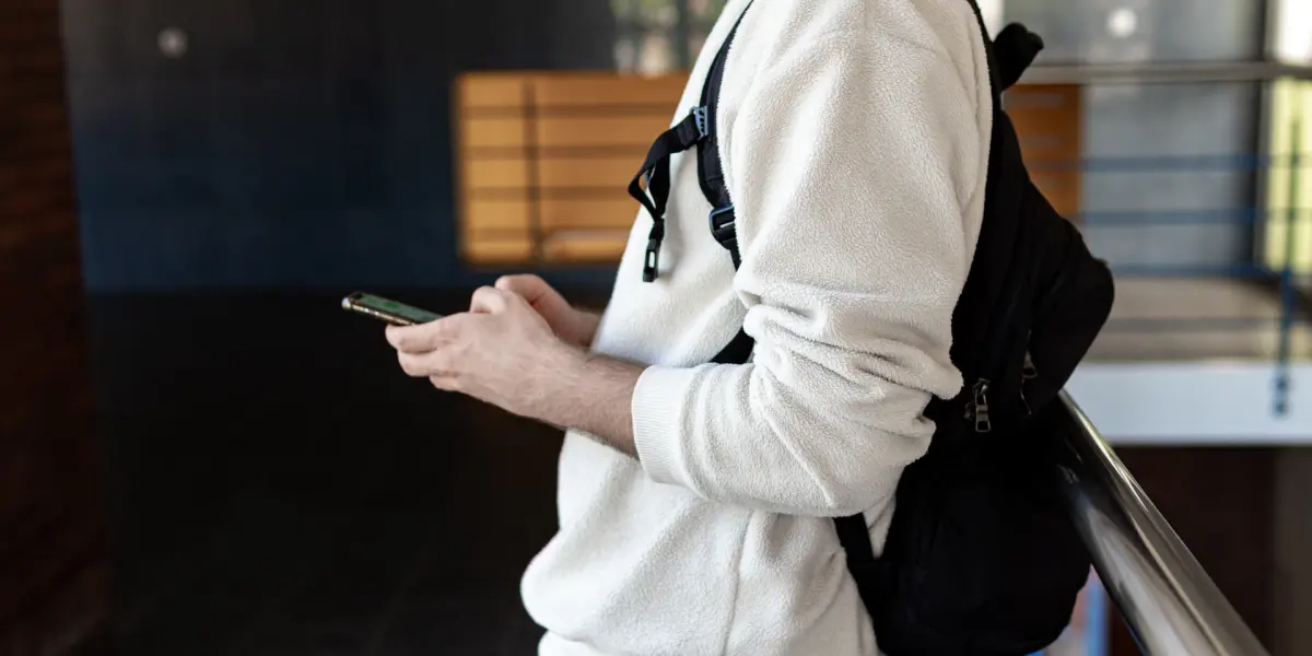 Person in a white jumper and backpack checking a phone by a railing, head out of frame